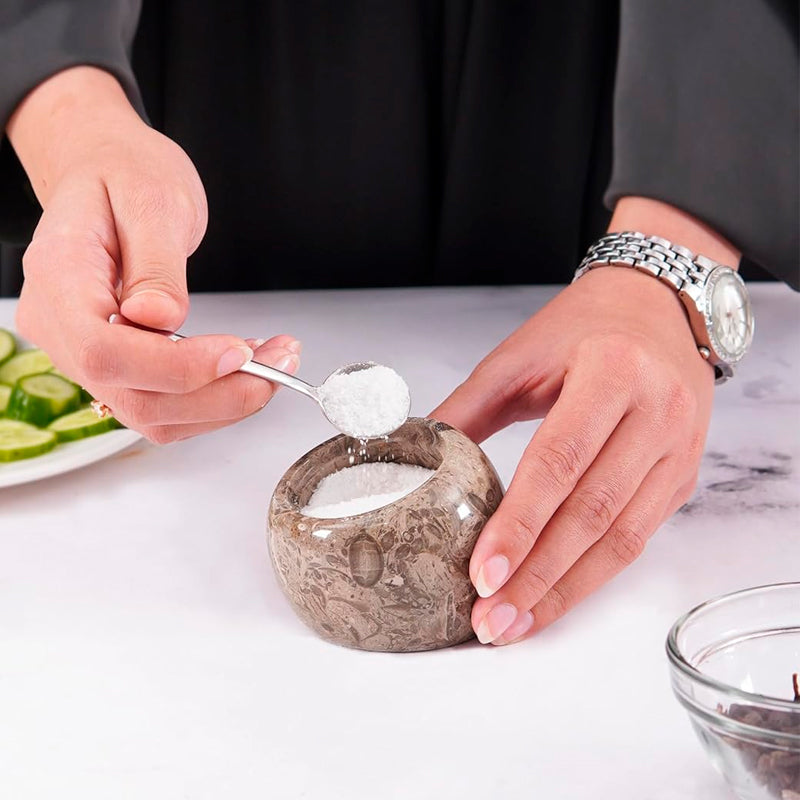 Person scooping salt from a stone container with a spoon on a white surface.
