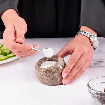 Person scooping salt from a stone container with a spoon on a white surface.
