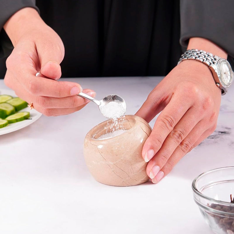 Person using a spoon to scoop out a small container of salt on a white surface.