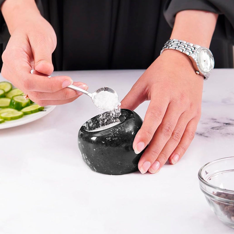 Person using a black stone mortar and pestle on a marble surface with a plate of sliced cucumbers in the background.