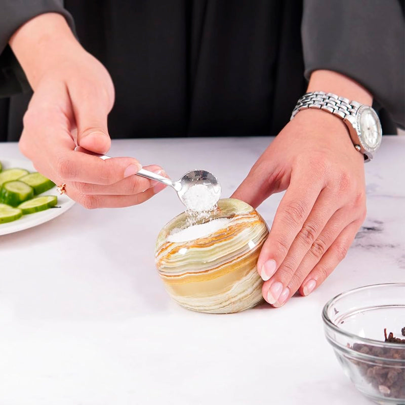 Person scooping a spoonful of salt from a decorative salt cellar on a white surface.