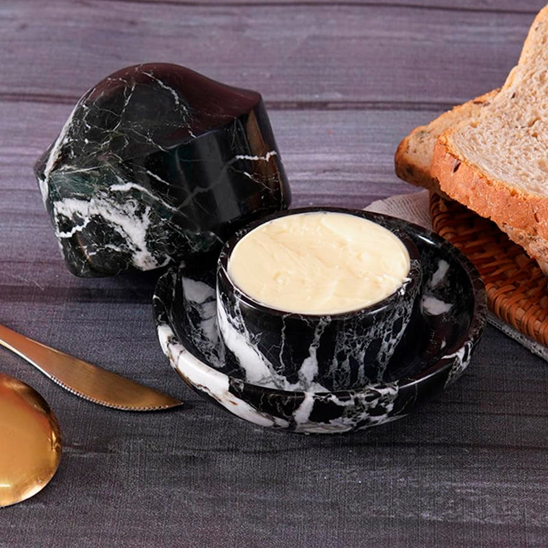Black marble butter dish with a pat of butter on a wooden surface with bread and a knife.