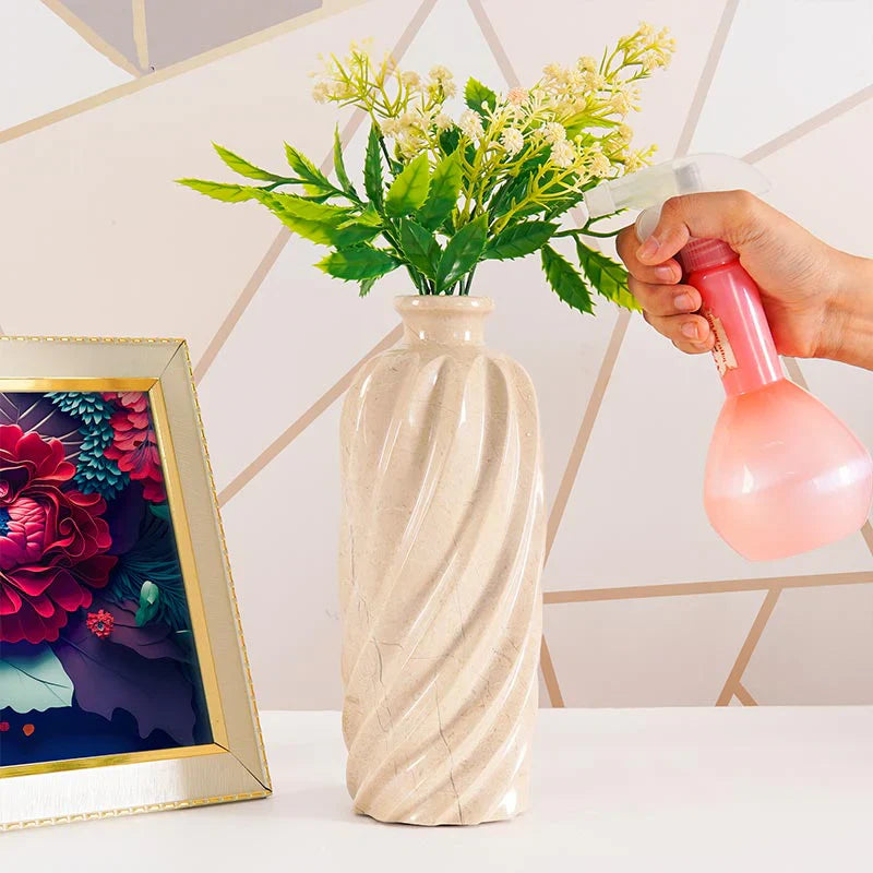 Person watering a vase with flowers on a table next to a framed picture.