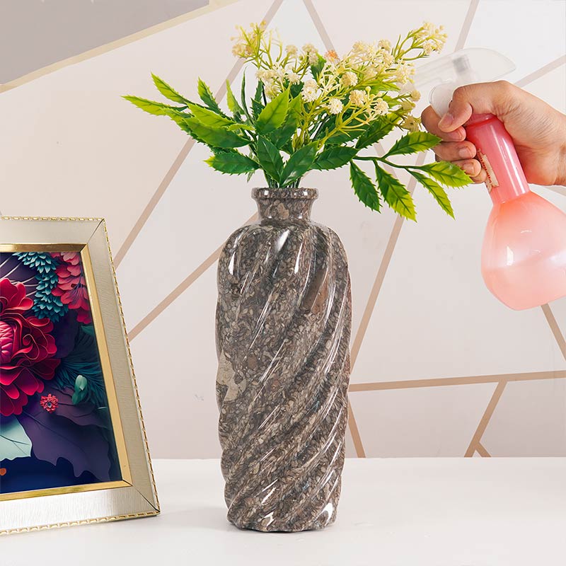 Person watering a vase with flowers on a table next to a framed picture.